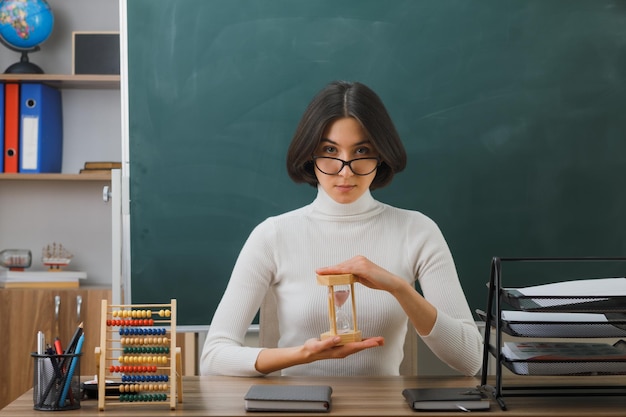 joven maestra segura de sí misma con gafas sosteniendo un reloj de arena sentada en el escritorio con herramientas escolares en el aula