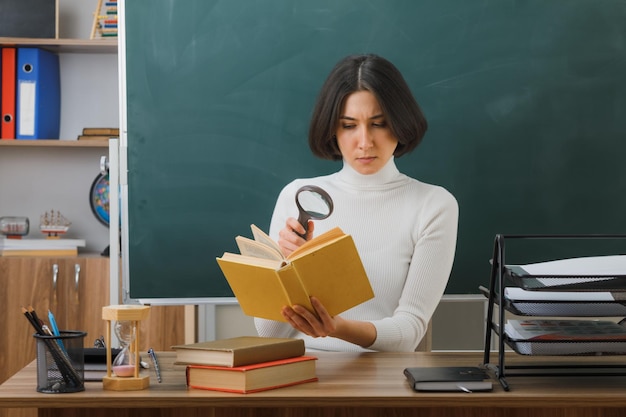 joven maestra segura de sí misma mirando un libro con lupa sentada en el escritorio con herramientas escolares en el aula