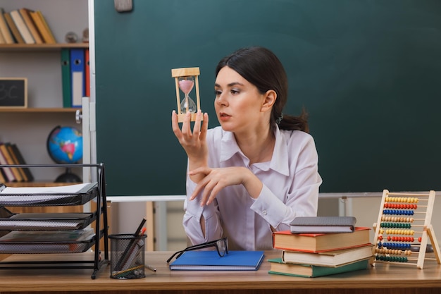 joven maestra segura de sí misma sosteniendo y mirando el reloj de arena sentada en el escritorio con herramientas escolares en el aula