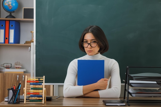 joven maestra segura de sí misma usando anteojos sosteniendo un cuaderno sentado en el escritorio con herramientas escolares en el aula