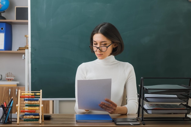 joven maestra segura de sí misma usando anteojos sosteniendo y mirando papel sentado en el escritorio con herramientas escolares en el aula