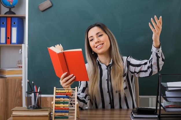 Foto joven maestra sentada en el escritorio de la escuela frente a la pizarra en el aula leyendo un libro preparándose para la lección feliz y emocionada