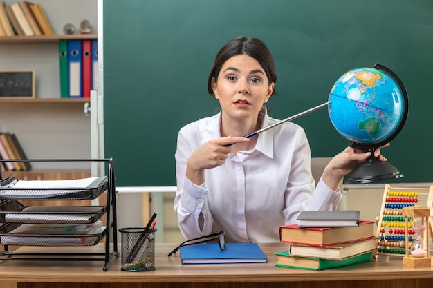 Joven maestra sentada a la mesa con herramientas escolares sosteniendo y poniendo el puntero en el globo en el aula