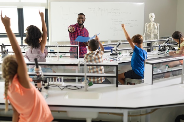 Joven maestro afroamericano interrogando a estudiantes primarios multirraciales con las manos levantadas