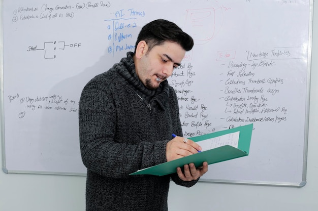 Un joven maestro está parado frente a la pizarra sosteniendo un archivo en la mano y comprobando.