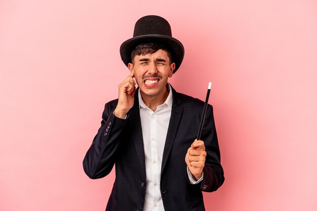 Joven mago caucásico sosteniendo una varita aislada en un fondo rosado cubriendo las orejas con las manos.