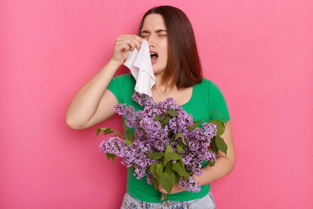 Una joven malsana con el pelo castaño y una camiseta verde informal sonándose la nariz con un pañuelo que sufre alergia a las flores lilas posando aislada sobre un fondo de pared rosa