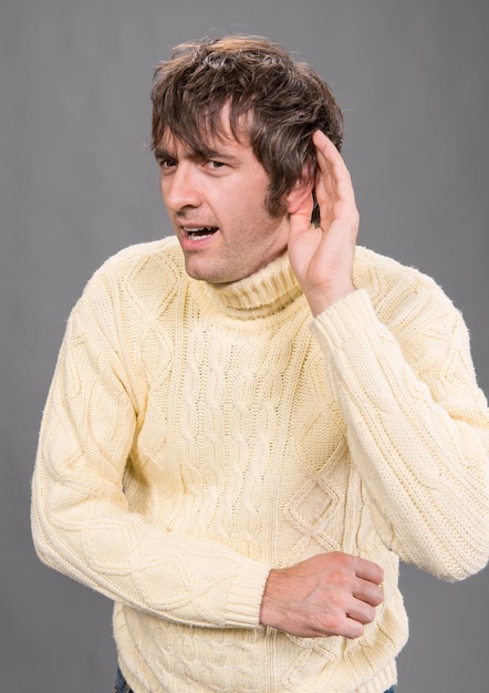 Joven con la mano detrás de la oreja escuchando atentamente contra un fondo gris