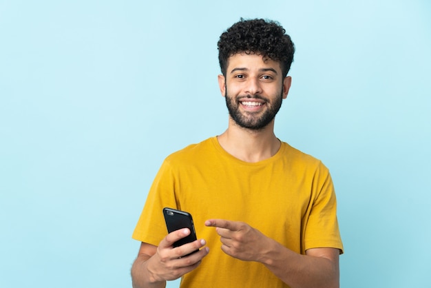 Joven marroquí aislado en azul mediante teléfono móvil y apuntando