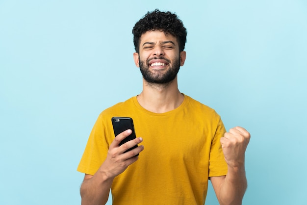 Joven marroquí aislado en azul mediante teléfono móvil y haciendo gesto de victoria