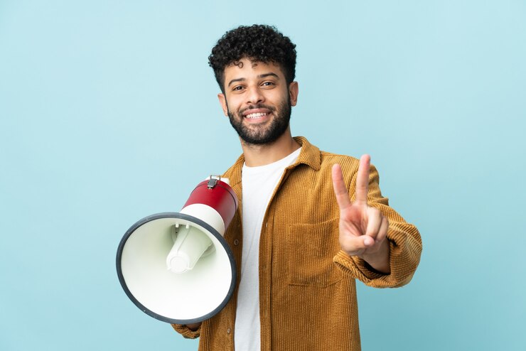 Foto joven marroquí aislado en la pared azul sosteniendo un megáfono y sonriendo y mostrando el signo de la victoria