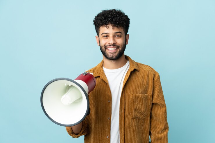 Foto joven marroquí aislado en la pared azul sosteniendo un megáfono y sonriendo mucho