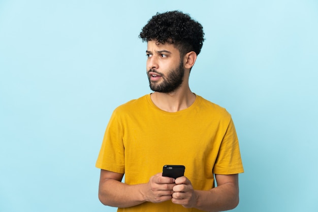 Joven marroquí aislado en la pared azul mediante teléfono móvil y mirando hacia arriba