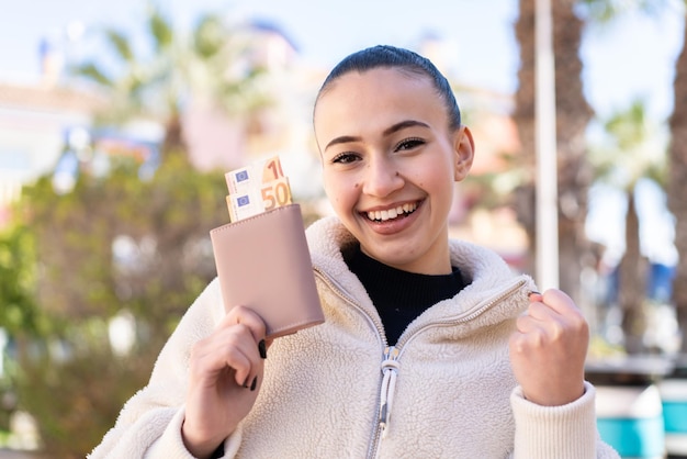 Joven marroquí al aire libre sosteniendo billetera con dinero y celebrando una victoria