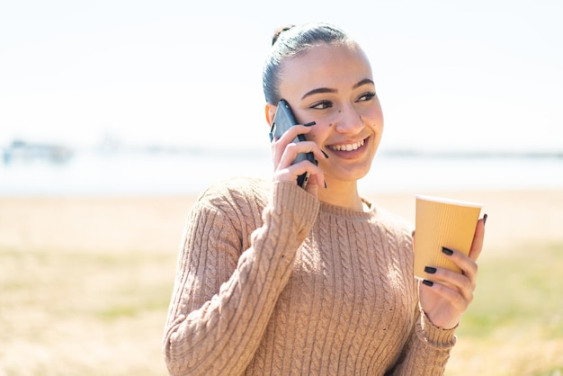 Joven marroquí al aire libre usando un teléfono móvil y sosteniendo un café con expresión feliz