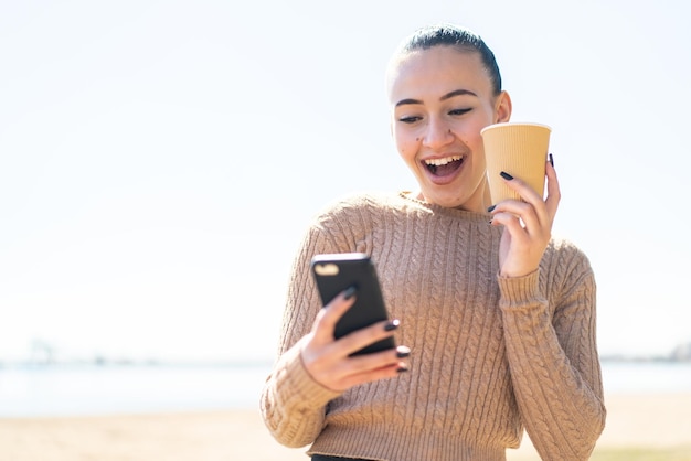 Joven marroquí al aire libre usando un teléfono móvil y sosteniendo un café con expresión feliz