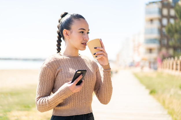 Joven marroquí al aire libre usando teléfono móvil y sosteniendo un café