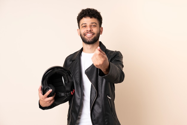 Joven marroquí con un casco de motocicleta aislado en pared beige haciendo gesto de dinero