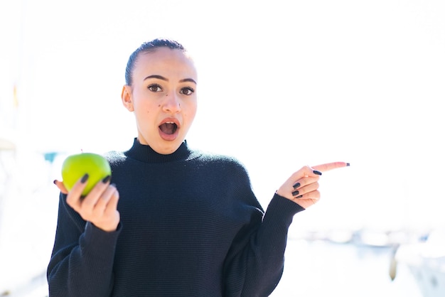 Joven marroquí con una manzana al aire libre sorprendida y apuntando hacia el lado