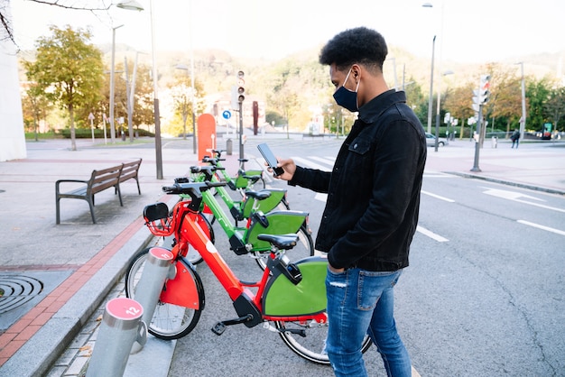 Joven marroquí que usa su teléfono móvil para recoger una bicicleta eléctrica alquilada en el parque de bicicletas de la calle y usa una mascarilla para la pandemia de coronavirus