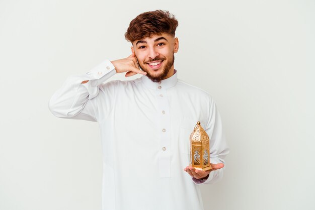 Joven marroquí con ropa típica árabe sosteniendo una lámpara de Ramadán aislada en blanco mostrando un gesto de llamada de teléfono móvil con los dedos.