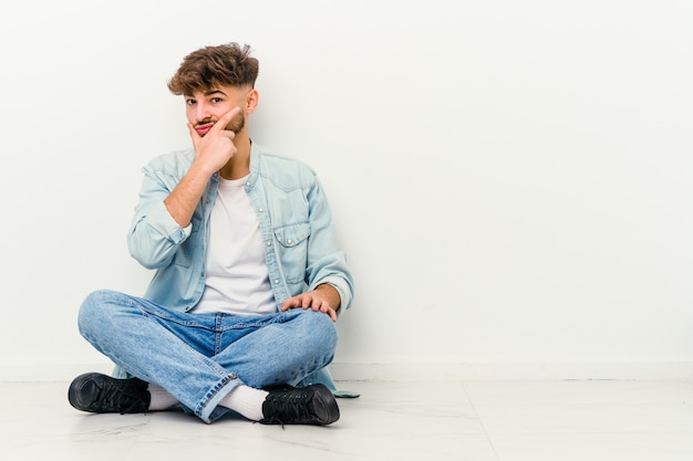 Joven marroquí sentado en el suelo aislado en la pared blanca contemplando, planificando una estrategia