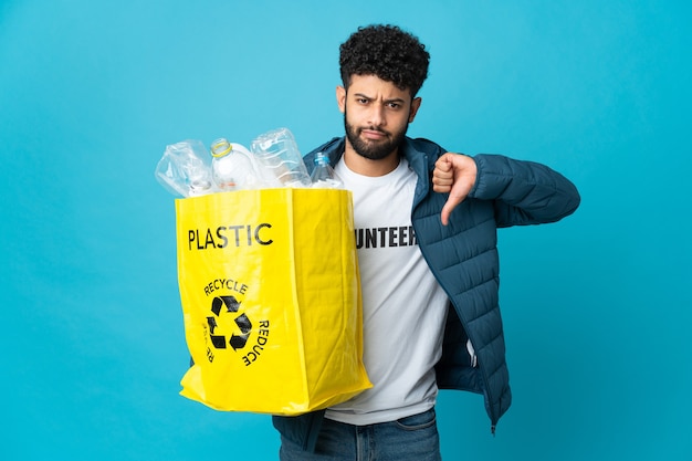 Joven marroquí sosteniendo una bolsa llena de botellas de plástico para reciclar mostrando el pulgar hacia abajo con expresión negativa