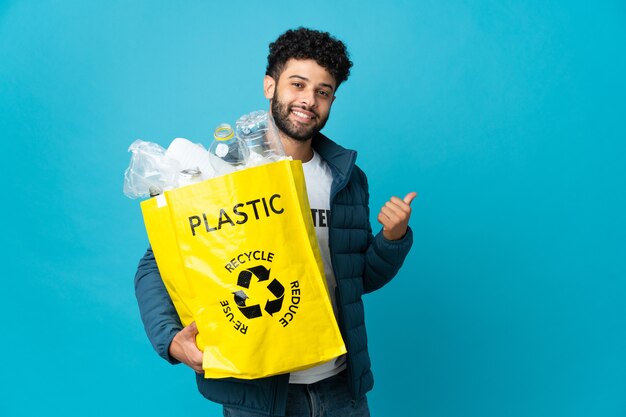 Joven marroquí sosteniendo una bolsa llena de botellas de plástico para reciclar sobre aislado apuntando hacia atrás