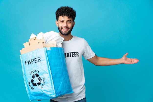 Joven marroquí sosteniendo una bolsa de reciclaje llena de papel para reciclar extendiendo las manos hacia el lado para invitar a venir