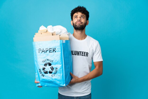 Joven marroquí sosteniendo una bolsa de reciclaje llena de papel para reciclar sobre pared aislada y mirando hacia arriba