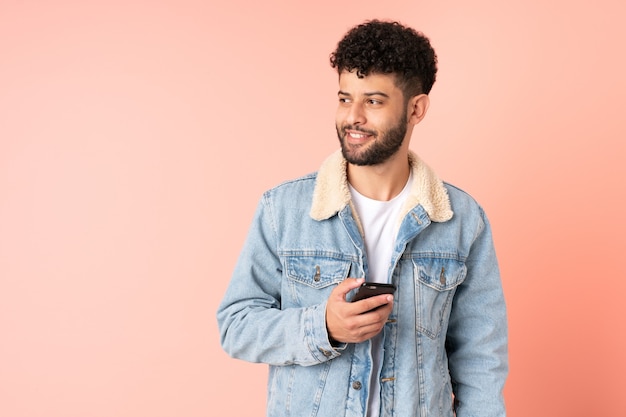 Joven marroquí mediante teléfono móvil aislado sobre fondo rosa mirando hacia el lado y sonriendo