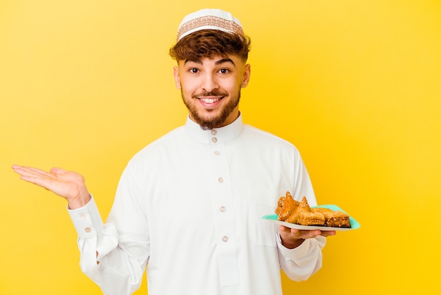 Joven marroquí vestido con el traje típico árabe comiendo dulces árabes aislado sobre fondo amarillo mostrando un espacio de copia en una palma y sosteniendo otra mano en la cintura.