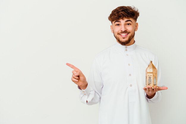 Joven marroquí vistiendo una ropa típica árabe sosteniendo una lámpara de Ramadán aislada en blanco sonriendo y apuntando a un lado, mostrando algo en el espacio en blanco.