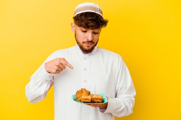 Joven marroquí vistiendo el traje típico árabe comiendo dulces árabes aislado en la pared amarilla