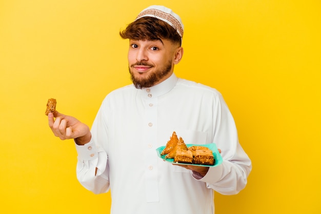 Joven marroquí vistiendo el traje típico árabe comiendo dulces árabes aislado sobre fondo amarillo