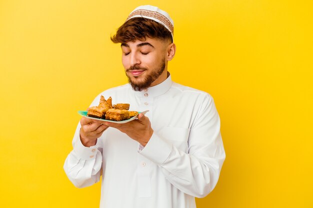 Joven marroquí vistiendo el traje típico árabe comiendo dulces árabes aislado sobre fondo amarillo