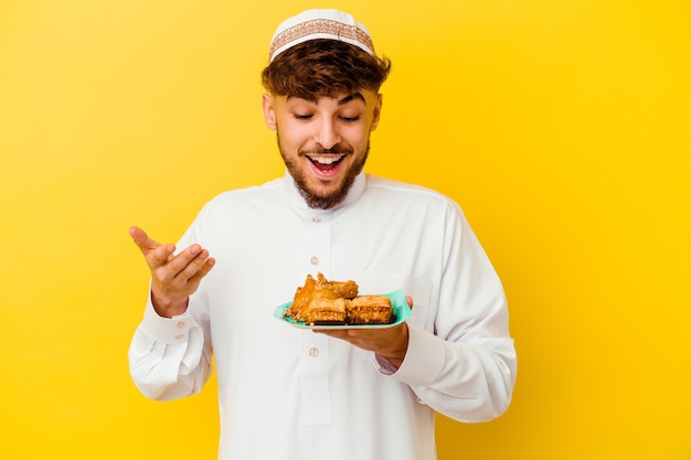 Joven marroquí vistiendo el traje típico árabe comiendo dulces árabes aislado sobre fondo amarillo