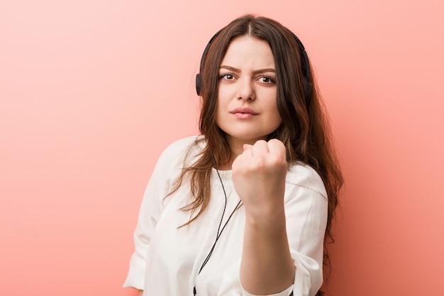 Joven más tamaño mujer con curvas escuchando música con auriculares mostrando puño, expresión facial agresiva.