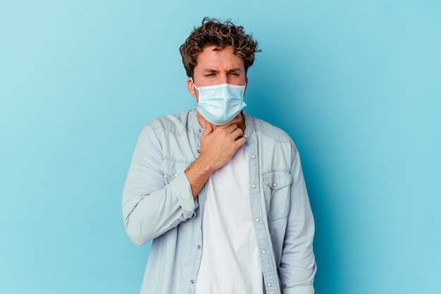 Joven con una máscara antiviral aislada en la pared azul sufre dolor de garganta debido a un virus o infección