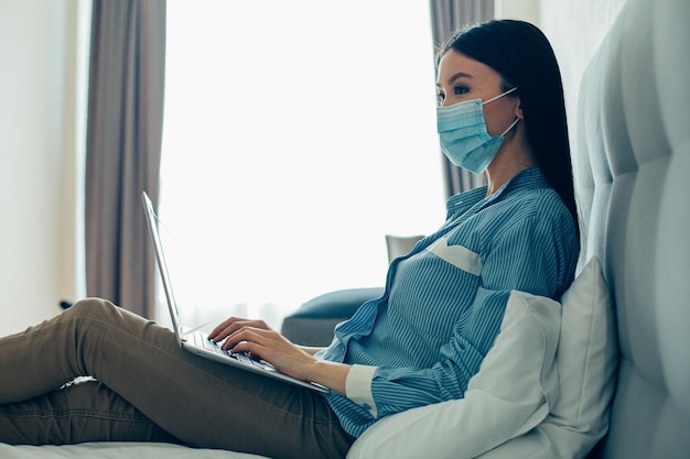 Joven con una máscara médica sentada en la cama con una computadora portátil moderna en el regazo