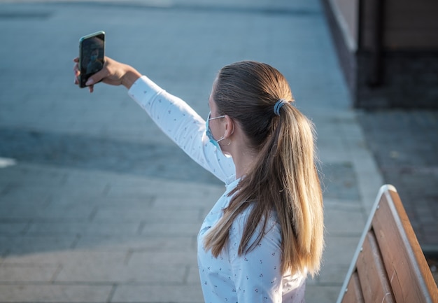 Una joven con una máscara médica se toma una selfie con su teléfono inteligente. modo máscara durante la pandemia de covid-19