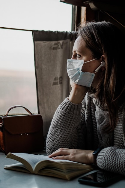 Una joven con una máscara médica viaja en tren durante una pandemia de coronavirus Tomando precauciones en el transporte público La niña está leyendo un libro y escuchando música en el tren