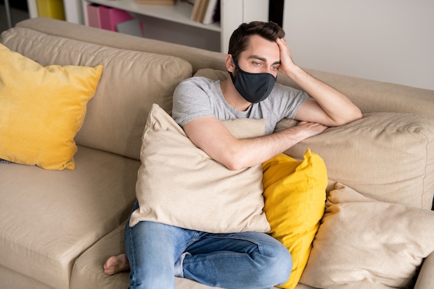 Joven con máscara protectora cansado del aislamiento en casa sentado con almohadas en el sofá y pensando en el futuro