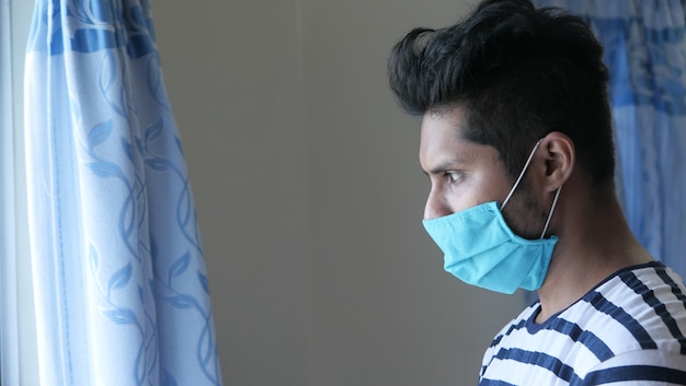 Un joven con máscara en su rostro mirando por la ventana