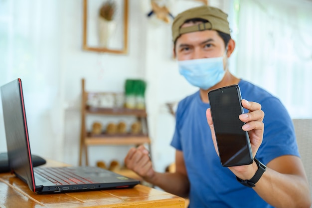 Joven con una máscara trabaja en línea con éxito Mostrar teléfono móvil con pantalla en blanco Hombre asiático que trabaja desde casa