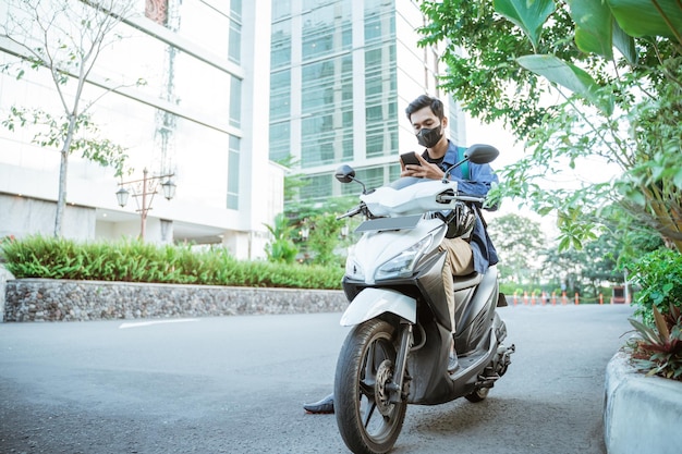 Joven con máscara usando un teléfono celular en una motocicleta en el fondo de la calle de la ciudad