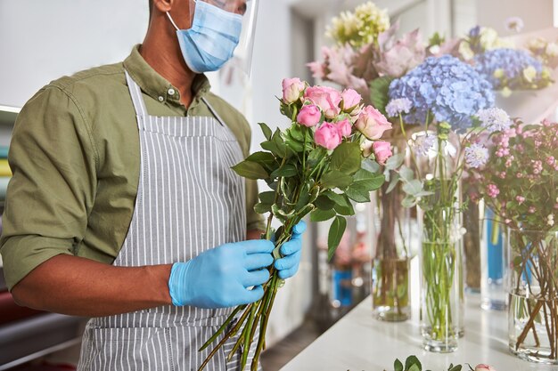 Joven con máscara y vidrio protector en floristería mientras hace ramos de flores