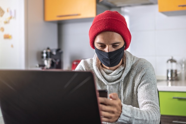 Un joven con una mascarilla negra está usando una computadora portátil y un teléfono mientras trabaja en casa