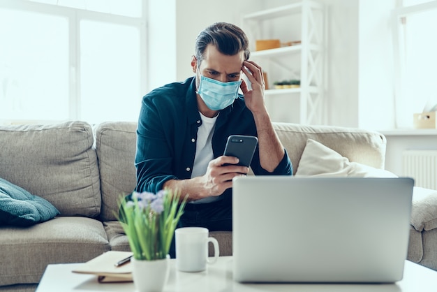 Joven en mascarilla protectora con teléfono inteligente mientras trabaja desde casa