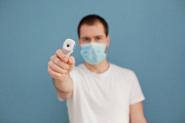 Un joven con mascarilla quirúrgica está usando un termómetro infrarrojo en un fondo azul